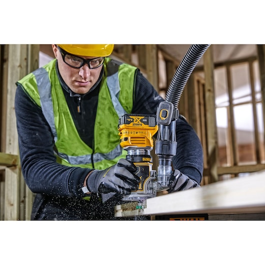 A person uses a DEWALT DCW604 router to shape wood in a construction setting, wearing safety gear and gloves.