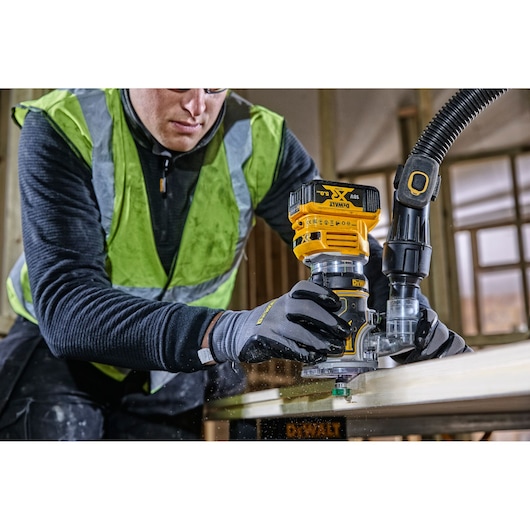 Worker uses a DEWALT DCW604 router on wood, wearing gloves and a safety vest in a workshop.