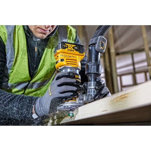 Worker using a DEWALT DCW604 cordless router on wood, wearing gloves and a safety vest in a workshop setting.