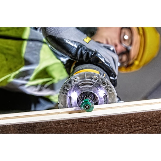 Close-up of DEWALT DCW604 router in use on wood, operator wearing gloves, helmet, and safety glasses.