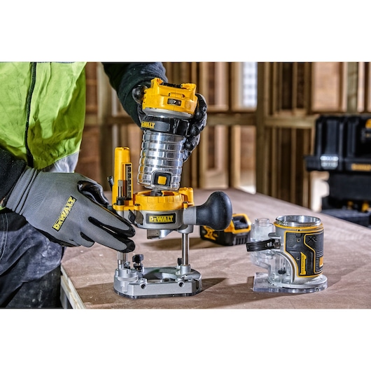 Person assembles DEWALT DCW604 router on workbench with extra accessory, wearing gloves in a workshop.