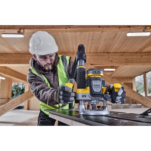 A person wearing safety gear operates a DEWALT XR cordless router (SKU: DCW620H2) on a wooden surface inside a timber structure.