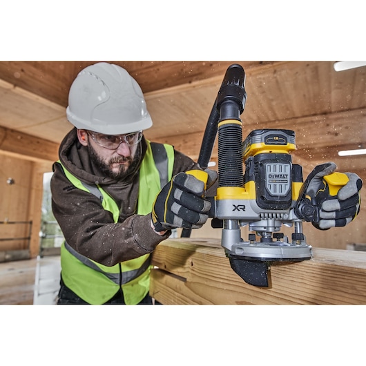 A worker wearing a safety vest, gloves, and a hard hat uses a DEWALT XR cordless router to shape the edge of a wooden plank in a construction setting. The router is connected to a dust extraction hose, and sawdust is visible in the air.