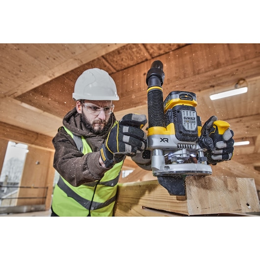 A construction worker wearing a safety helmet and high-visibility vest is operating a DEWALT XR router (model DCW620H2) on a wooden beam inside a timber building. Wood shavings are visible as the tool is in use.