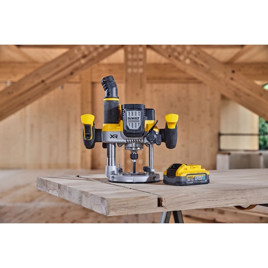 A DEWALT cordless plunge router with two black and yellow handles and an attached dust extraction port is placed on a wooden workbench inside a wooden structure. Next to the router is a DEWALT 18V POWERSTACK battery pack.