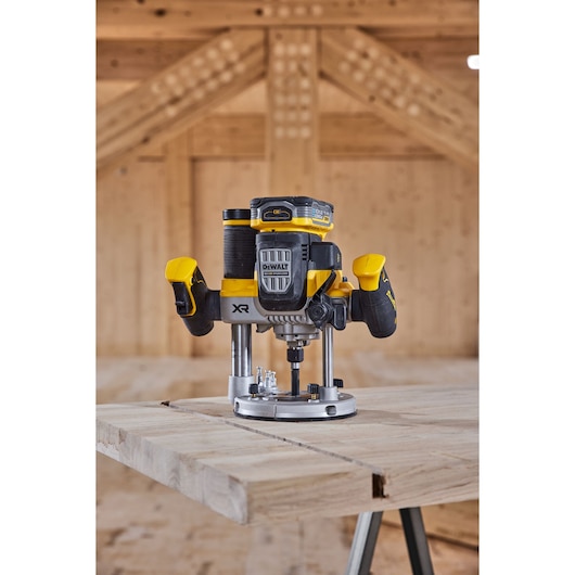 A DEWALT cordless router tool, model DCW620H2, is placed on a wooden workbench inside a wooden workshop. The tool is yellow and black with dual handles and a battery pack on top.