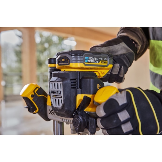 Close-up of a person using a DEWALT cordless power tool with POWERSTACK 18V battery, wearing protective gloves and high visibility clothing, in a workshop or construction environment.