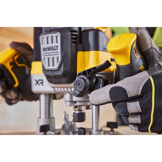 Close-up view of a DEWALT XR cordless router being adjusted by a gloved hand. The router is yellow and black, with visible branding and control levers.