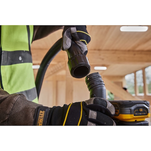 Close-up of a person wearing gloves connecting hoses on a DEWALT power tool in a wooden interior construction setting.