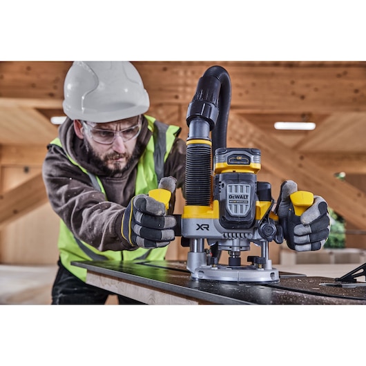 A worker wearing protective gear operates a DEWALT DCW620NT power tool, likely a router, on a piece of wood in an indoor construction setting.