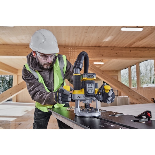 A person wearing safety equipment operates a DEWALT DCW620NT XR cordless router on a wooden work surface in a workshop with wooden beams and windows.