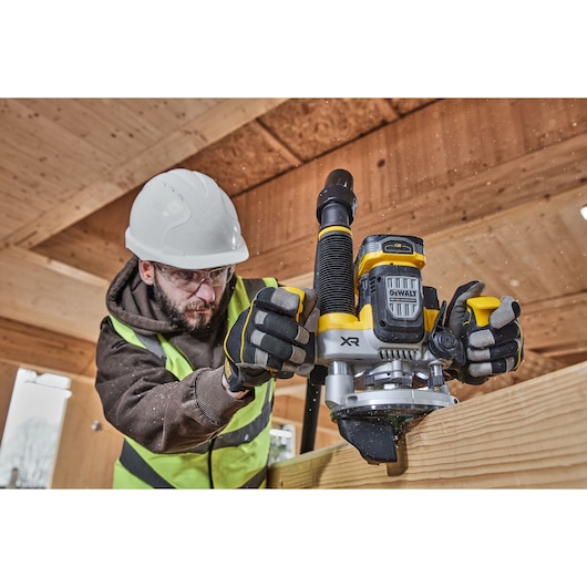 A person wearing a safety helmet, high-visibility vest, and work gloves is operating a DEWALT XR cordless router (DCW620NT) on a wooden beam in a construction setting. The face of the individual is obscured for privacy.