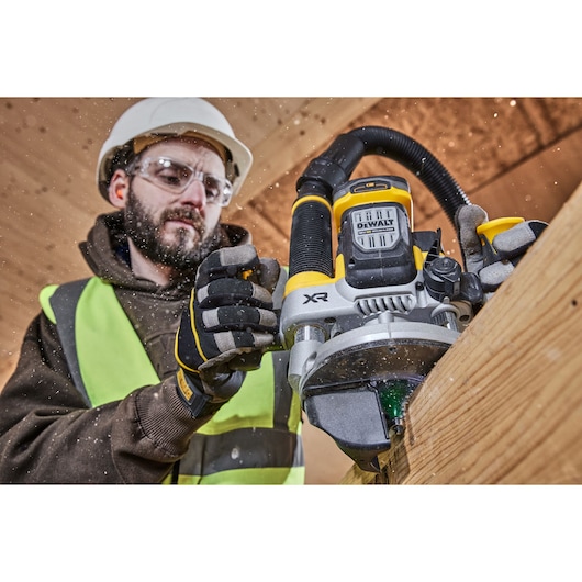 A worker using a DEWALT DCW620NT cordless router to shape a wooden beam. The worker is wearing safety gear including a helmet, gloves, and a high-visibility vest. Wood shavings are visible in the air.