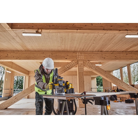 A worker wearing safety gear operates a DEWALT power tool, specifically a router, on a large piece of wood in a timber-framed construction site. The worker is standing at a workbench, surrounded by wooden beams and natural light.