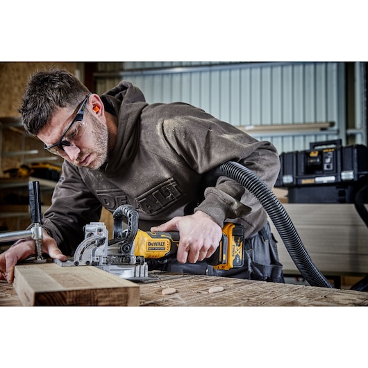 A person using a DEWALT DCW682N cordless jointer on a piece of wood in a workshop setting. The tool is yellow and black, connected to a dust extraction hose, and clamped to a wooden workbench.