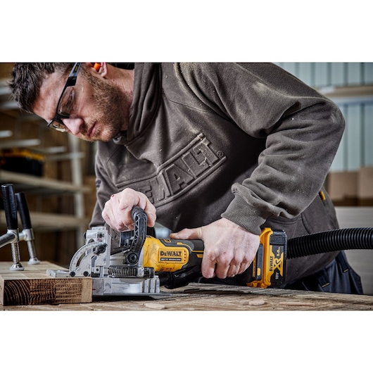 A person using a DEWALT cordless joiner tool (model DCW682N) to work on a piece of wood in a workshop. The tool has a yellow and black design and is connected to a dust extraction hose.