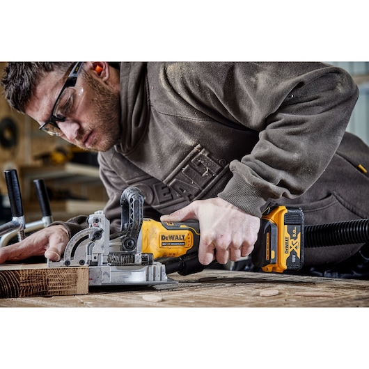 A person is using a DEWALT cordless biscuit joiner tool to work on a piece of wood in a workshop. The tool is yellow and black with a DEWALT XR 18V battery attached.