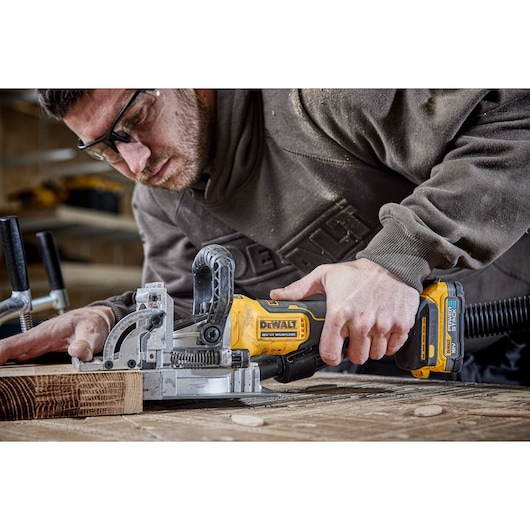 A person using a DEWALT cordless biscuit joiner (model DCW682N) to cut slots into a piece of wood in a workshop setting.