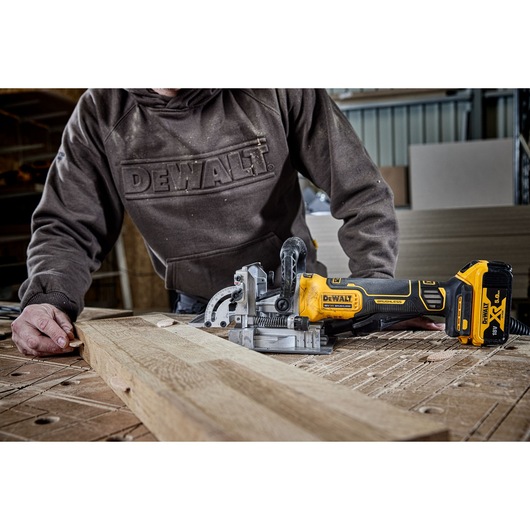 A person using a DEWALT cordless joiner tool to cut a piece of wood on a workbench in a workshop environment. The person is wearing a brown DEWALT hoodie and the tool is marked with 18V XR and DEWALT branding.