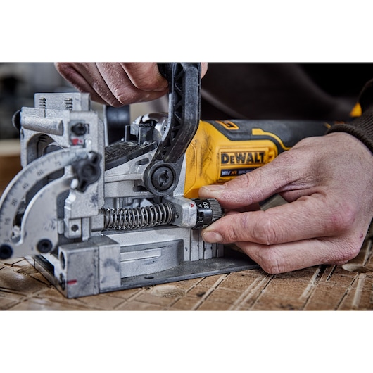 Close-up of a person adjusting a DEWALT DCW682P2 power tool, specifically a joiner, on a wooden surface. The hands are working on the adjustment knob and handle, with visible detail on the tool's metal components.