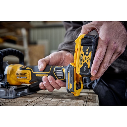 Close-up of a person inserting an 18V 5.0Ah XR lithium-ion battery into a DEWALT cordless tool, placed on a wooden surface in a workshop setting.
