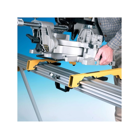 A close-up of a DEWALT miter saw mounted on a sturdy metal saw stand with yellow brackets. A person is adjusting or securing the saw onto the stand. The background is blue.