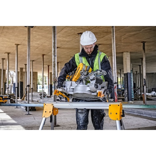 A construction worker wearing a high-visibility vest and hard hat is operating a DEWALT sliding compound miter saw mounted on a DEWALT saw stand at a building site.