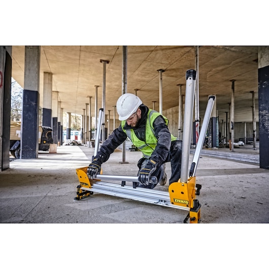 A construction worker wearing a safety helmet, high-visibility vest, and gloves is setting up a DEWALT DE7033 work support stand at an indoor construction site. The worker is kneeling on the concrete floor surrounded by support columns and unfinished building structures.