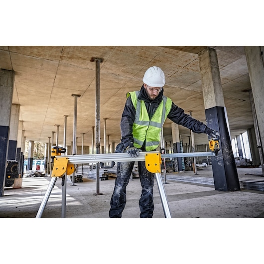 A worker wearing a safety helmet and a high-visibility vest is adjusting a DEWALT DE7033 work support stand at a construction site with concrete columns and exposed ceiling.