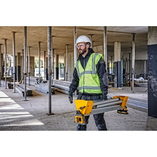 A construction worker wearing a high-visibility vest and hard hat carries a DEWALT DE7033 tool stand inside a building under construction. The background shows metal support columns and building materials.