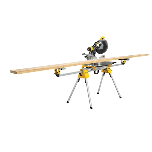 A DEWALT mitre saw mounted on a yellow and silver stand, cutting a long wooden board. The saw is positioned in the center, with adjustable extensions on both sides to support the board. The background is white.