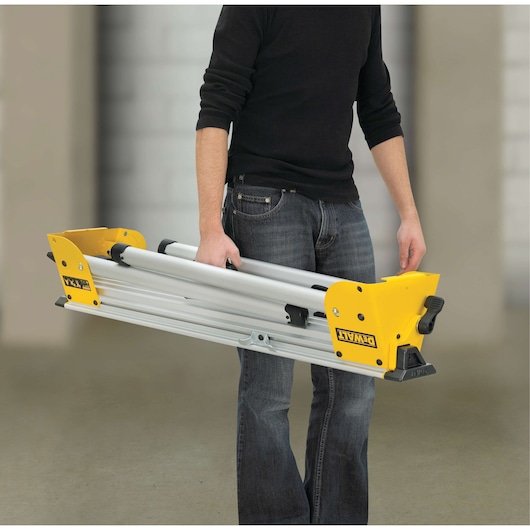 A person carrying a folded DEWALT DE7033 aluminum stand with yellow end pieces. The product is compact and portable, shown against an indoor background.