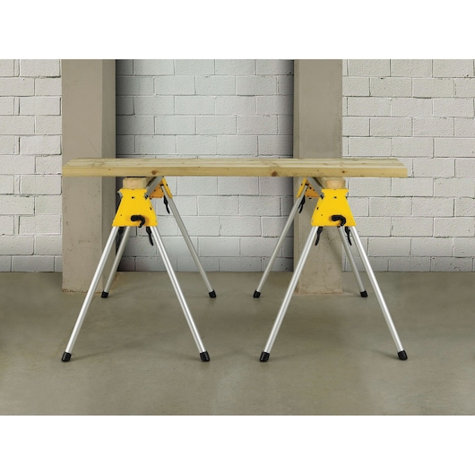 A pair of DEWALT DE7035 adjustable metal sawhorses with yellow and silver coloring, supporting a wooden plank in an indoor workshop setting.