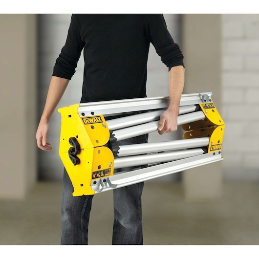 A person is carrying a folded DEWALT DE7035 stand with yellow and silver metal parts.