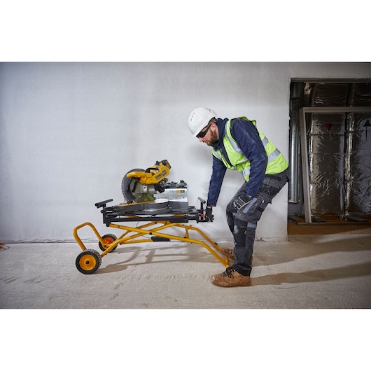 A person wearing a safety helmet, high-visibility vest, and workwear is handling a yellow DEWALT miter saw stand (model DE7260) with a mounted miter saw inside a construction environment. The stand has wheels for easy transport.
