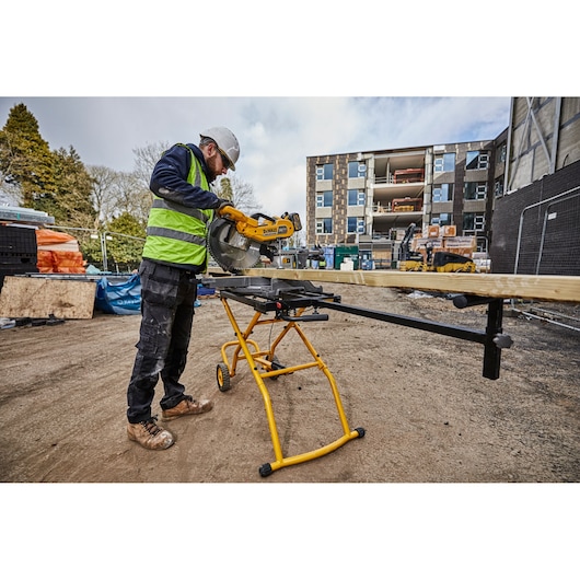 A construction worker uses a DEWALT miter saw mounted on a DEWALT DE7260 stand to cut a long piece of wood at an outdoor jobsite. The worker is wearing a safety helmet, high-visibility vest, and work boots. The background shows a building under construction with materials and equipment on site.