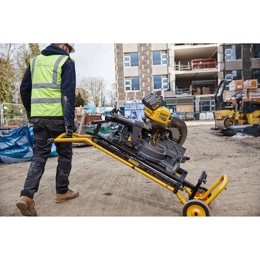 A construction worker transports a DEWALT miter saw attached to a yellow rolling stand at a building site. Construction materials and machinery are visible in the background.