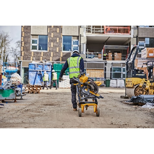 A construction worker wearing a high-visibility vest and helmet is pushing a DEWALT miter saw mounted on a mobile stand (model DE7260) across a busy construction site. The background shows other workers, building materials, and equipment in an active work zone.
