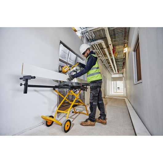 A worker wearing safety gear cuts a long white board with a sliding mitre saw mounted on a yellow DEWALT stand (model DE7260) in a construction hallway.