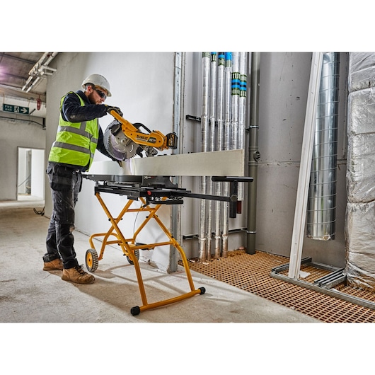 A person wearing safety gear operates a DEWALT miter saw mounted on a yellow DEWALT stand (model DE7260), cutting a long board indoors at a construction site.