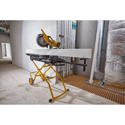 A DEWALT mitre saw mounted on a yellow mobile stand is set up indoors at a construction site, with a large white board positioned on the stand for cutting.