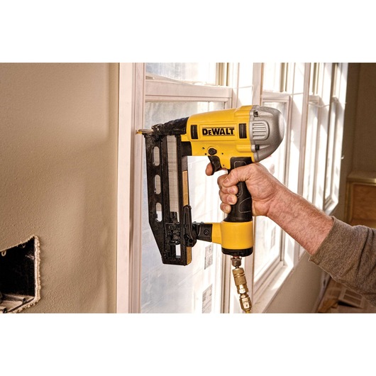 A person using a yellow and black DEWALT pneumatic nail gun to attach trim to a window frame inside a room.