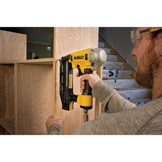 A person is using a DEWALT pneumatic nailer to attach wooden panels during a construction or carpentry project. The tool is yellow and black and the workspace appears to be indoors with unfinished wooden surfaces.