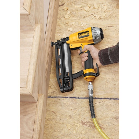 A DEWALT pneumatic nail gun (model DPN1664) being used to attach wooden stair trim on a plywood subfloor. The tool is connected to an air hose.