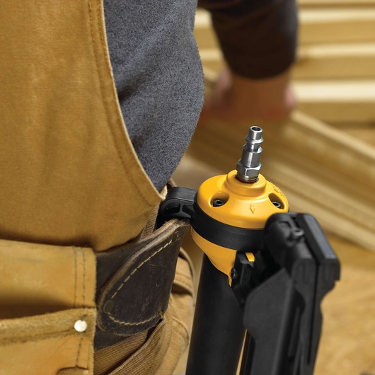 Close-up of a DEWALT pneumatic nailer tool attached to a worker's tool belt. The worker is wearing work overalls and standing near wooden planks.