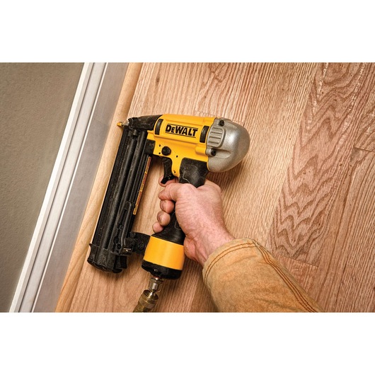 A person holding a DEWALT pneumatic nail gun, model DPN1850PP, using it to attach trim to a wooden floor.