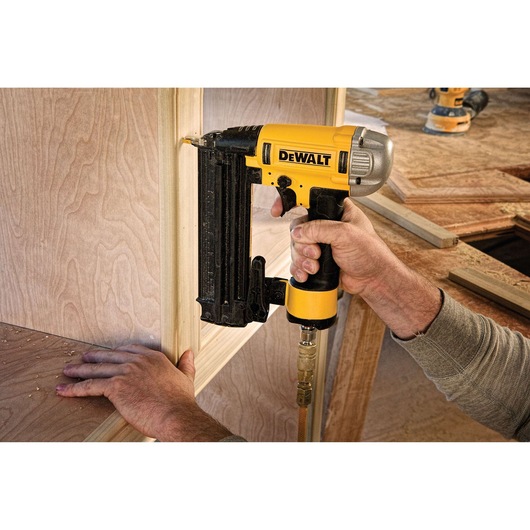 A person using a DEWALT pneumatic nailer to attach wooden trim to a wooden structure. The nailer is yellow and black and connected to an air hose. Workshop tools and wood are visible in the background.