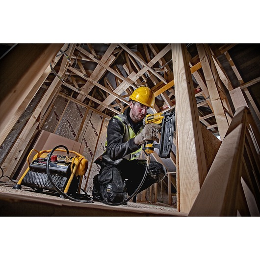 A person wearing a yellow hard hat and safety vest is working on a wooden construction site, using a DEWALT pneumatic nail gun connected to a DEWALT air compressor. The person is crouched among wooden beams, and their face is blurred.
