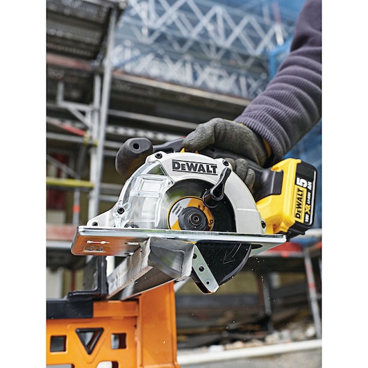 A DEWALT cordless circular saw (DT1910) is being used to cut through a metal piece, held by a person wearing a glove. The saw is positioned on an orange workbench, with construction scaffolding visible in the background.
