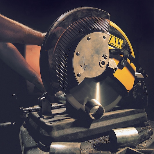 A DEWALT metal chop saw in use, cutting through a metal pipe. The tool is mounted on a workbench, surrounded by metal shavings and other metal pieces. The yellow brand logo is visible on the saw.